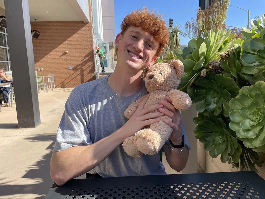 A person sitting at an outdoor café table, smiling and hugging a soft brown teddy bear in bright sunlight.