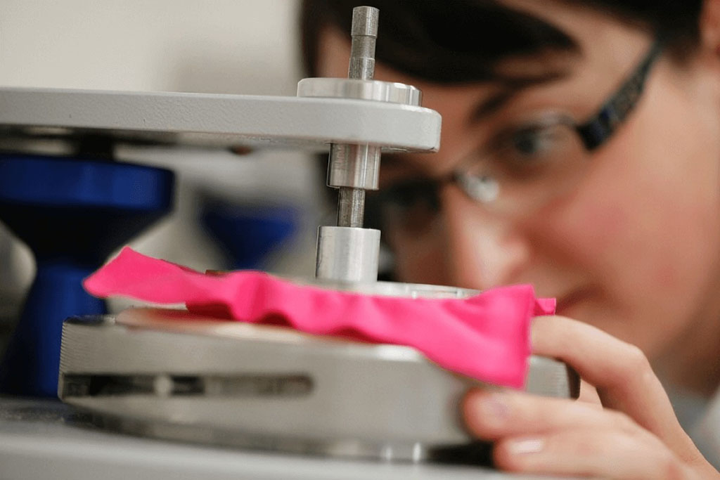 Technician conducting a fabric strength or abrasion resistance test using a laboratory machine with a pink textile sample under pressure for quality control.