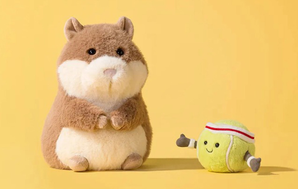 Cute Jellycat plush hamster sitting beside a smiling tennis ball plush with a headband on a yellow background.