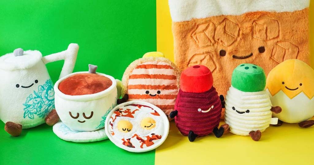 A colorful set of smiling food plush toys including a teapot, teacup, toast, and various breakfast items displayed on a bright green and yellow background.