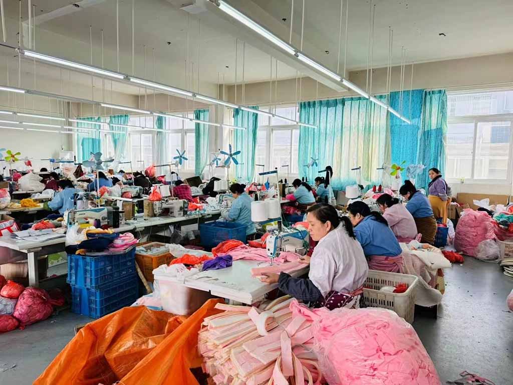 Workers sewing plush toys at factory tables surrounded by colorful fabric and stuffing materials.