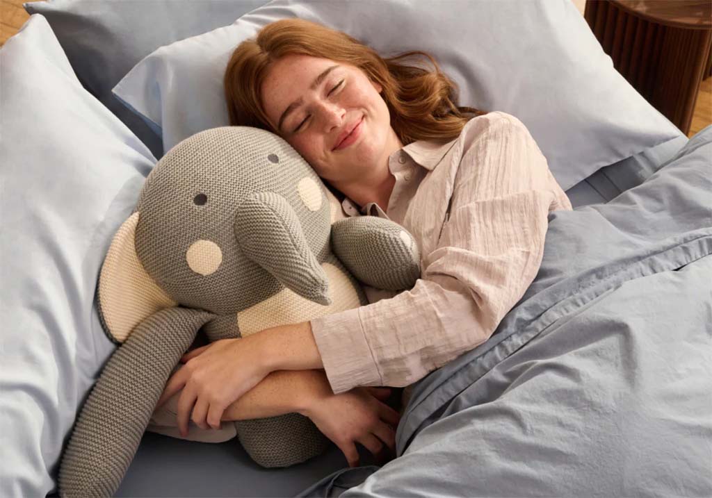 A woman peacefully sleeping in bed while hugging a large gray and cream elephant plush toy, wrapped in soft blue bedding.