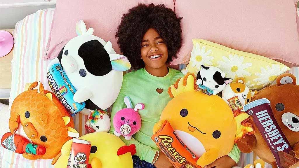 Smiling woman lying on a bed surrounded by colorful plush toys shaped like animals holding snack-themed accessories such as Pringles, Reese’s, and Hershey’s, with pastel pillows and bedding.