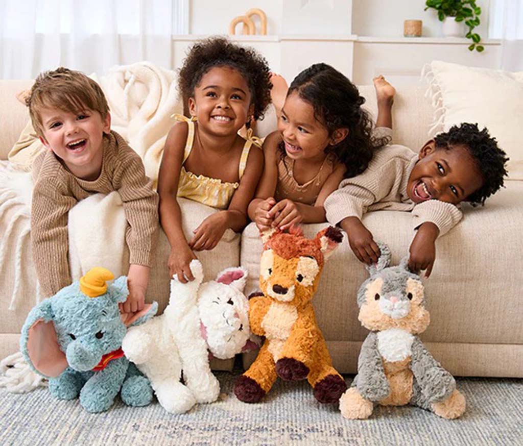 Four smiling children lying on a sofa and playing with soft plush toys, including an elephant, bunny, deer, and squirrel, in a cozy and bright living room setting.