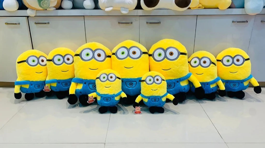 A lineup of bright yellow Minion plush toys in various sizes, all wearing blue overalls and goggles, displayed indoors on a tiled floor.