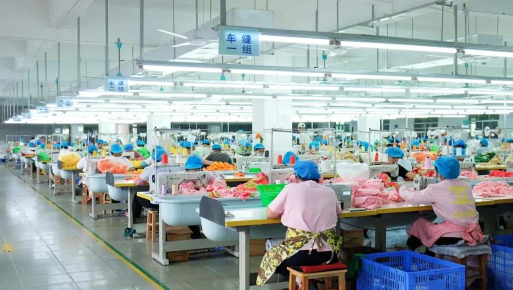 Factory workers in uniforms and hair covers sewing colorful plush toy fabrics in a clean, brightly lit sewing department.