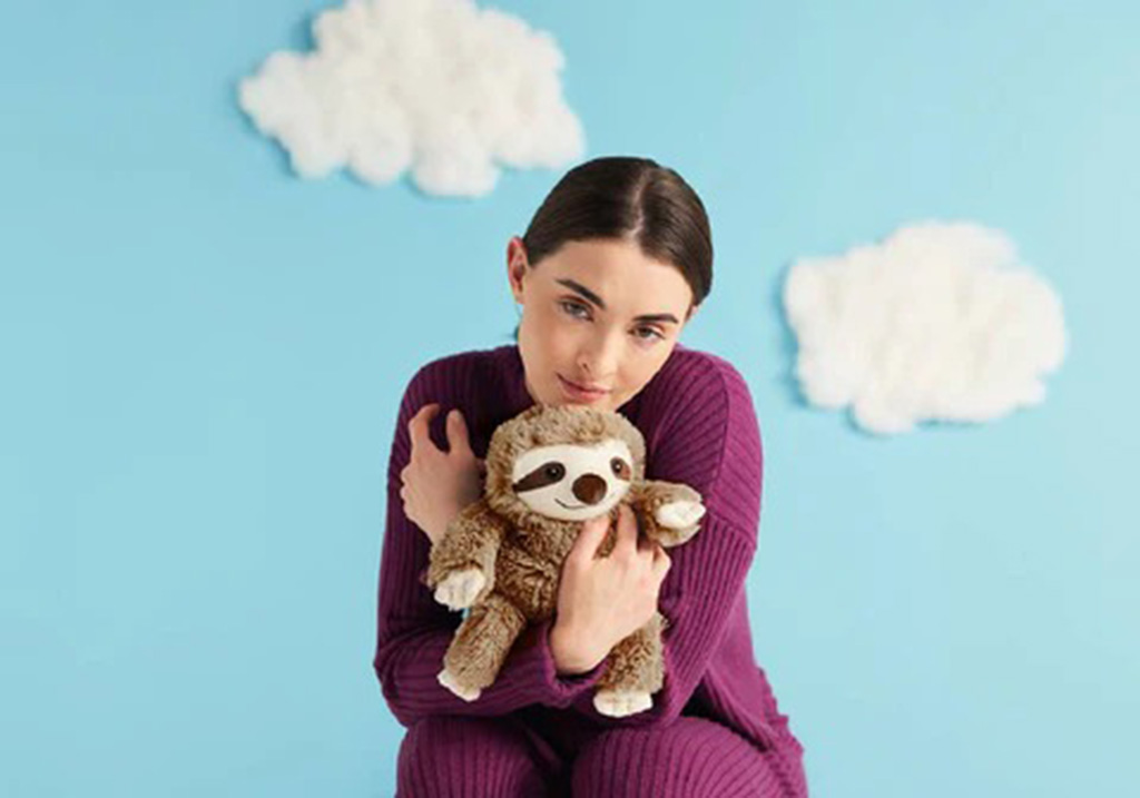 A woman in a purple outfit sitting against a blue background with fluffy white clouds, gently hugging a brown sloth plush toy for comfort.