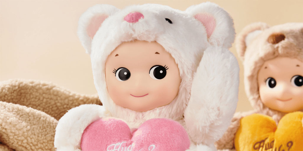 Adorable doll wearing a white bear costume and holding a pink heart-shaped pillow, with another doll in a brown bear outfit holding a yellow heart in the background.
