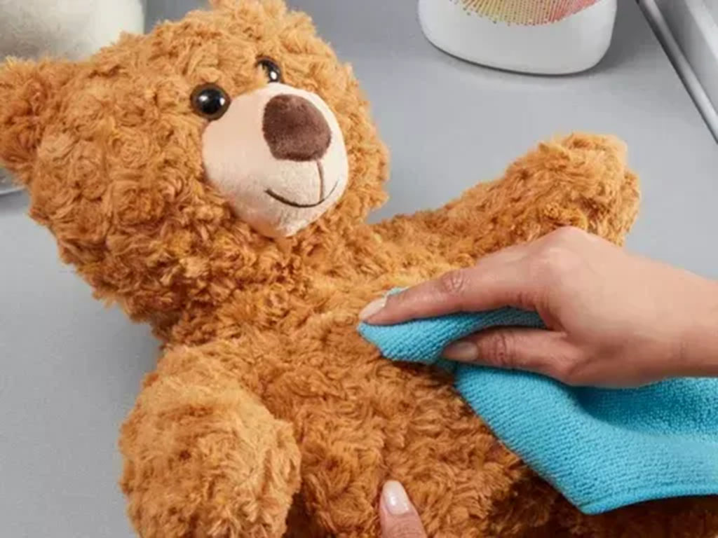 Person using a blue microfiber cloth to gently clean the surface of a brown teddy bear for light stain removal and care.