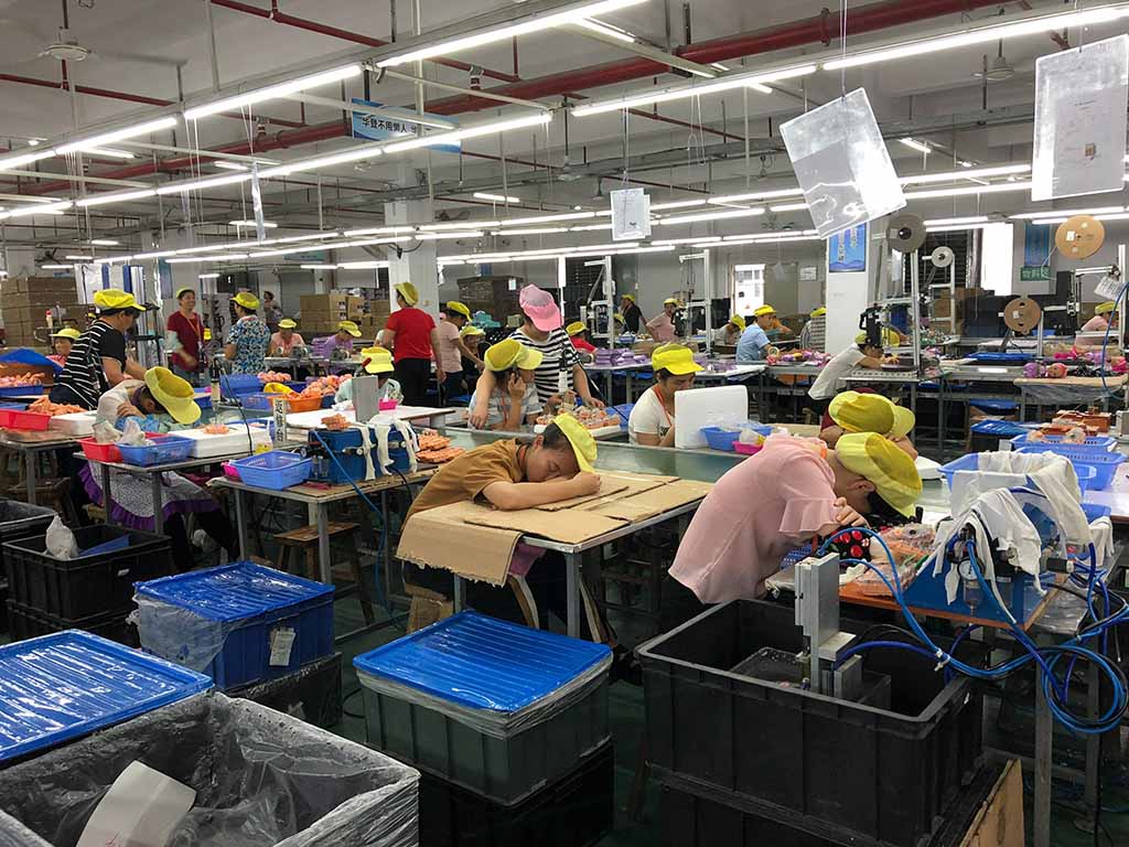 Factory workers wearing yellow caps assembling plush toys at organized workstations inside a well-lit production facility.