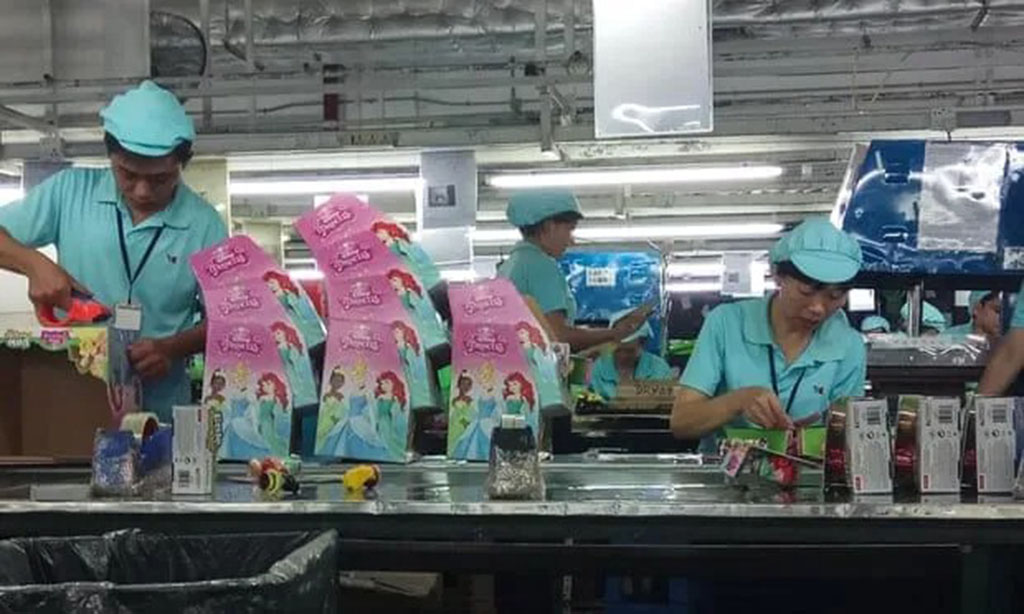 Factory workers in blue uniforms assemble and package colorful toy boxes on a production line under bright overhead lighting.