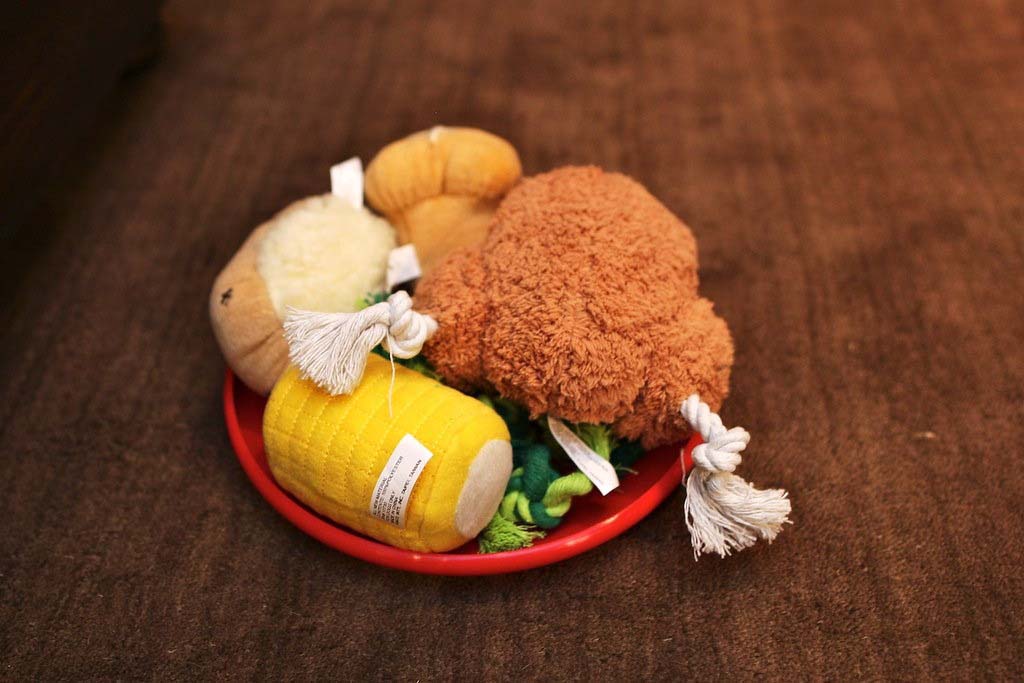 A red bowl filled with plush food toys, including a stuffed drumstick, corn, and bread, placed on a brown fabric surface.