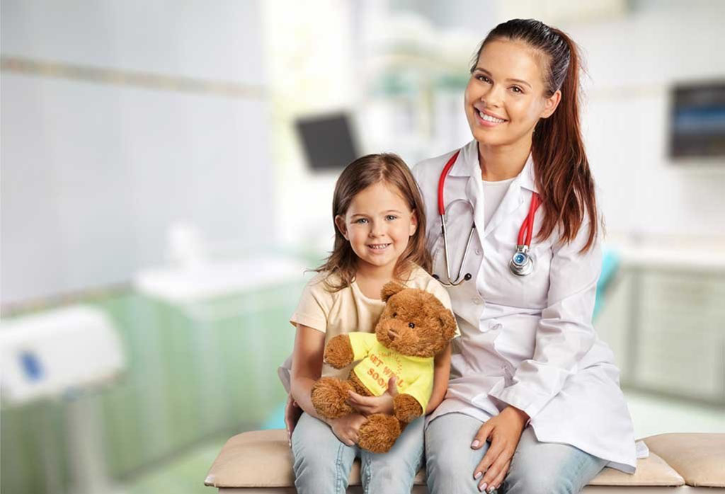 Female doctor with stethoscope sitting beside a young girl holding a brown teddy bear in a medical clinic, both smiling warmly.