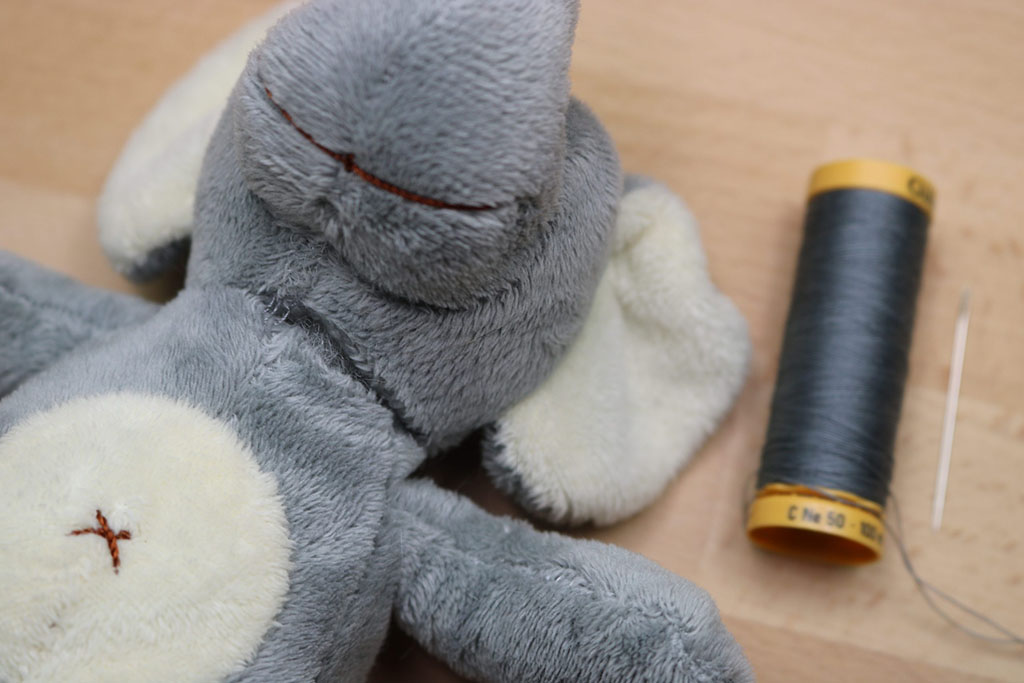 Grey plush toy with visible stitches beside a spool of thread and needle, showing the process of repairing a soft toy by hand sewing.