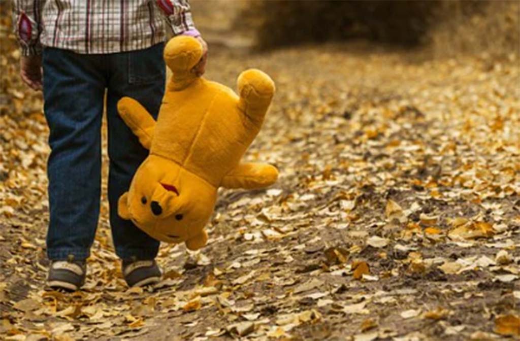 Child in jeans and sneakers holding an upside-down teddy bear while walking along a leaf-covered path in a forest during autumn.