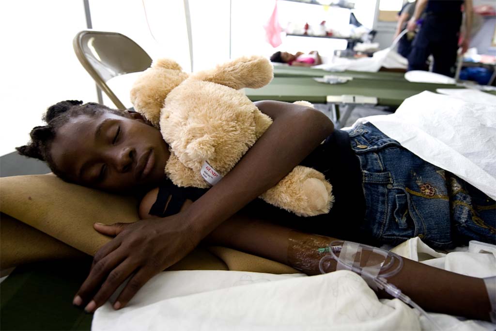 Young patient lying on a hospital bed with an IV line, peacefully holding a soft beige teddy bear for comfort.
