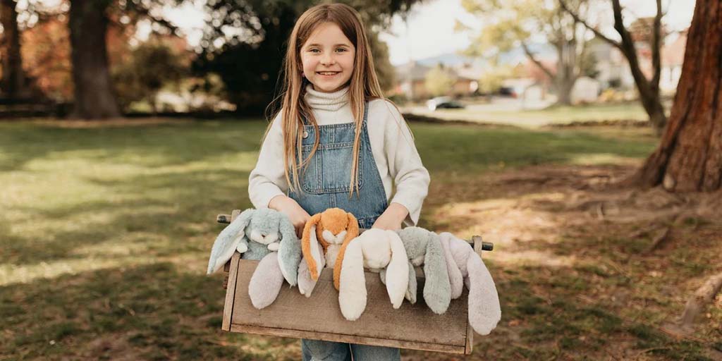 Smiling girl outdoors holding a wooden box filled with soft bunny plush toys in pastel colors.