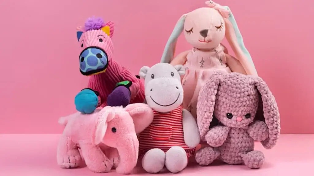 Group of colorful plush toys including a horse, hippo, elephant, and two bunnies sitting together against a pink backdrop.