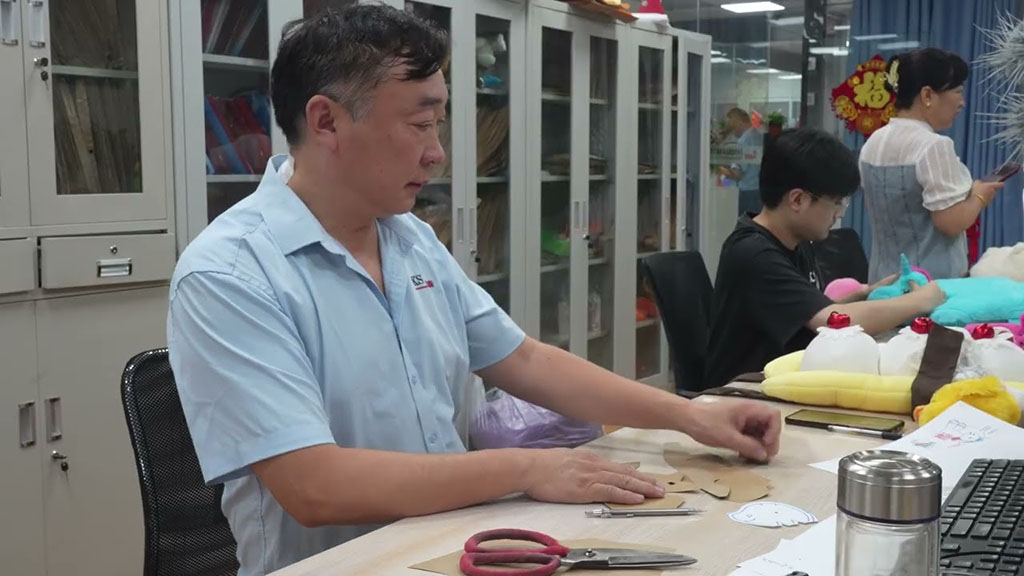 Designer working on plush toy pattern pieces at a table in a studio with other team members and colorful stuffed prototypes in the background.