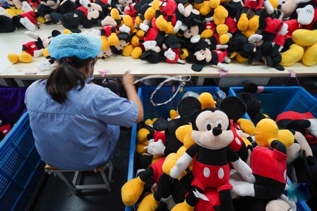 Factory worker wearing blue uniform and cap inspecting piles of Mickey Mouse plush toys during quality control and sorting process.