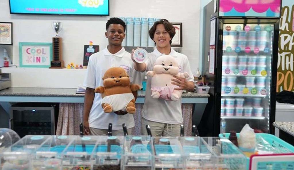 Two young men standing behind the counter of a dessert shop, smiling and holding cute baby-themed plush toys, with colorful containers and shop decor in the background.