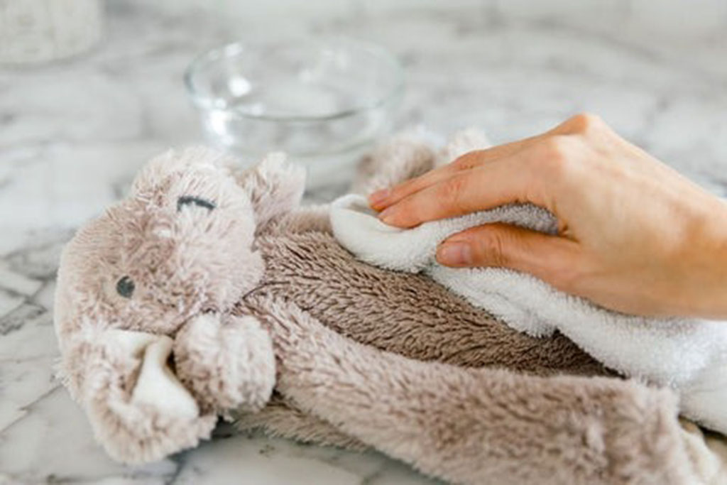 Hand gently wiping a soft brown stuffed animal with a white cloth on a marble surface, demonstrating surface cleaning for plush toys.