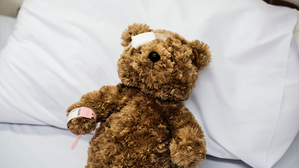 A brown teddy bear with a bandage on its head and a hospital wristband lies on a white pillow, symbolizing toy repair or care.