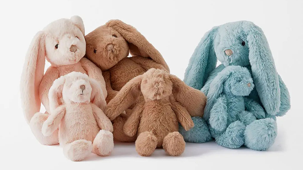 Group of plush bunny toys in pastel colors—cream, brown, and blue—sitting together, each with long floppy ears and soft fur, arranged in small and large sizes.