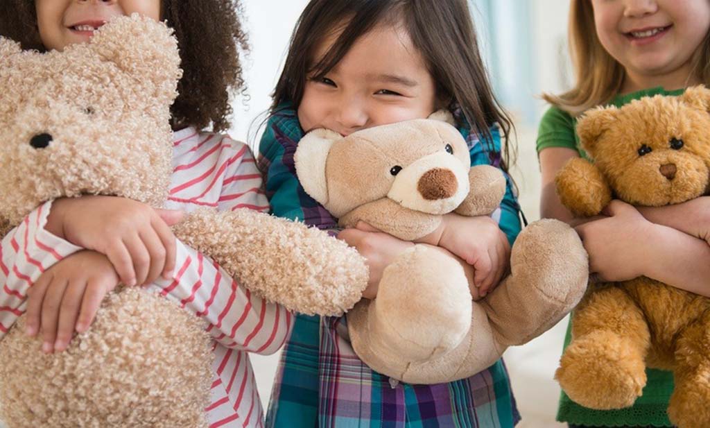 Three smiling children standing side by side, each hugging a soft teddy bear of different colors and sizes.