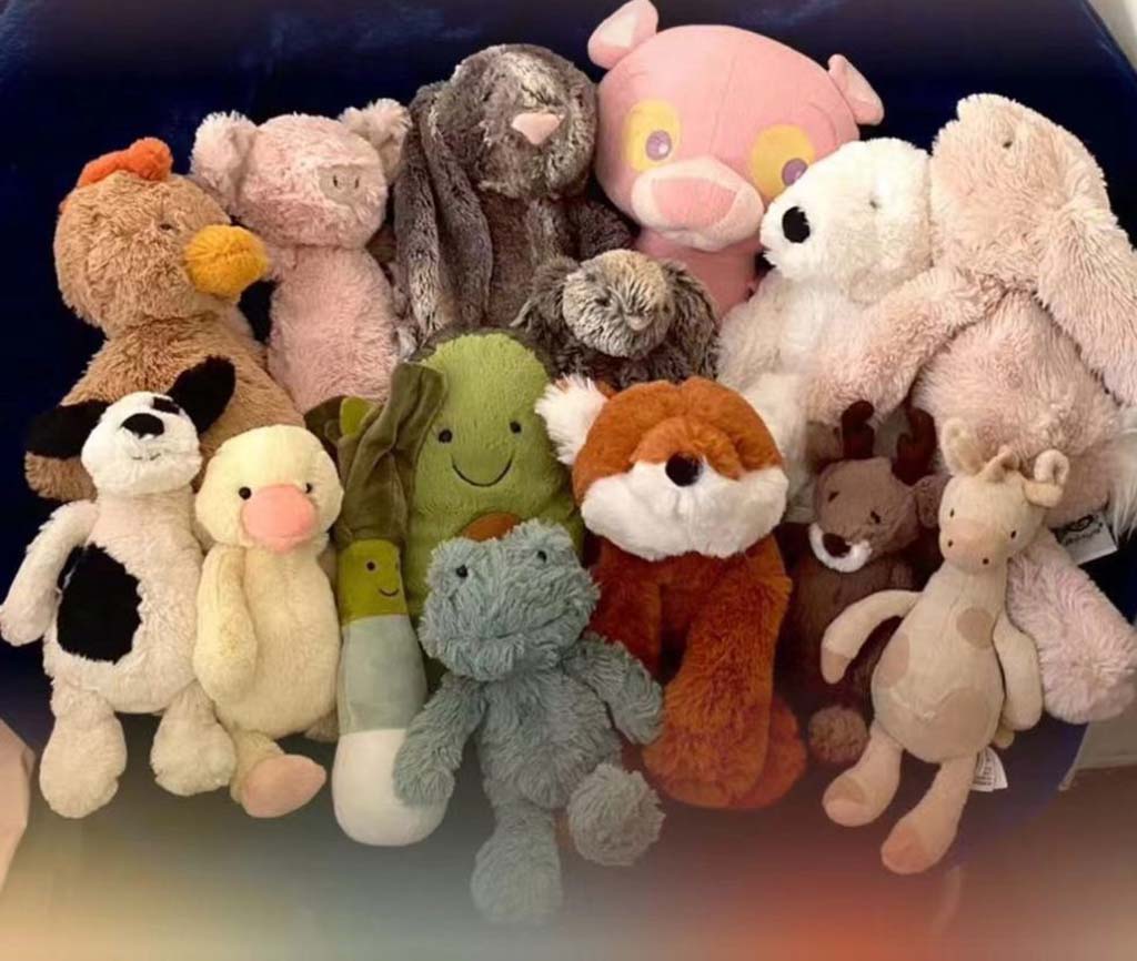 A cozy group of assorted plush toys including a fox, bunny, pig, duck, giraffe, and other soft animals arranged together on a dark background.