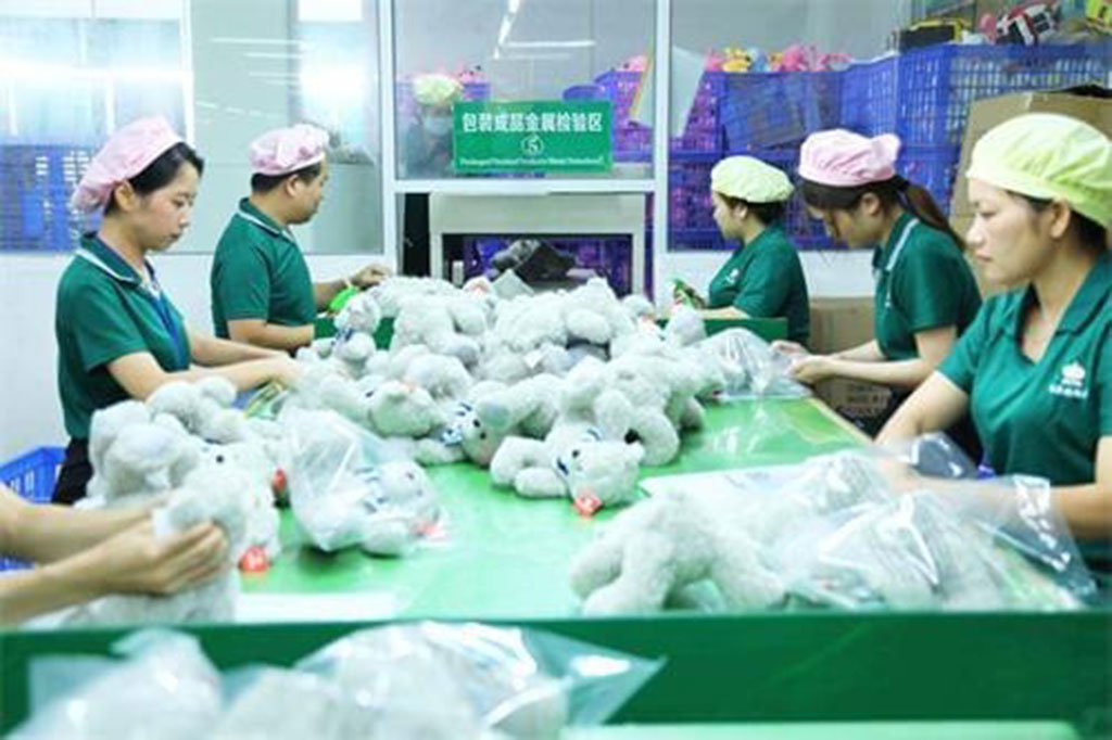 Factory workers in green uniforms and caps package soft plush toys into plastic bags on a green assembly table.
