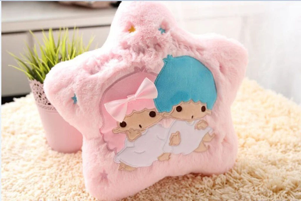 Soft pink star-shaped plush pillow featuring two cute cartoon characters with pastel blue and pink hair, placed on a fluffy carpet beside a potted plant.