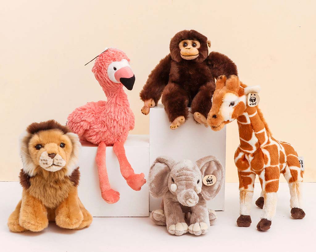A group of realistic animal plush toys including a lion, flamingo, monkey, giraffe, and elephant displayed together on white blocks against a beige background.