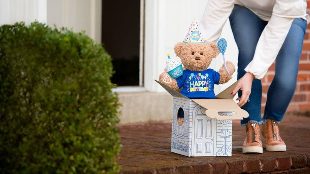 A woman places a birthday-themed teddy bear in a gift box on a doorstep, featuring a plush bear wearing a party hat and “Happy Birthday” shirt, symbolizing a surprise delivery.
