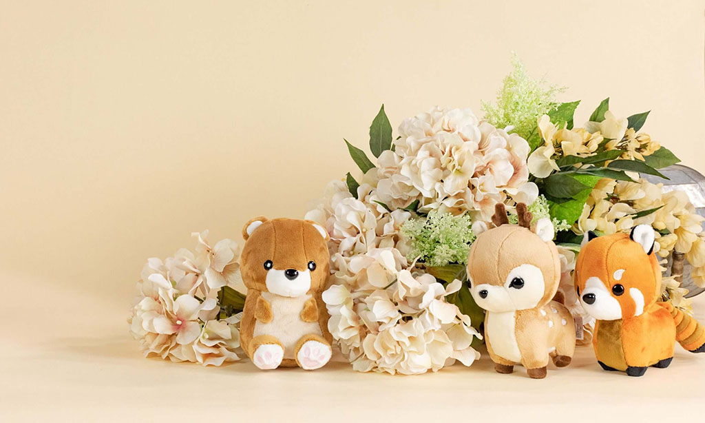 Three small plush toys—a bear, a deer, and a red panda—displayed in front of cream-colored hydrangea flowers on a beige background.