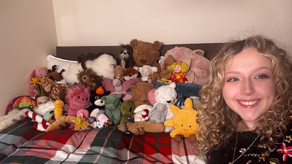 Smiling person with curly hair posing beside a large collection of colorful plush toys arranged on a bed.