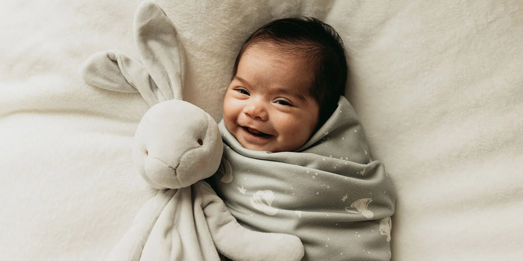 Smiling baby wrapped in a gray blanket cuddling a soft gray bunny plush toy on a white fluffy surface.