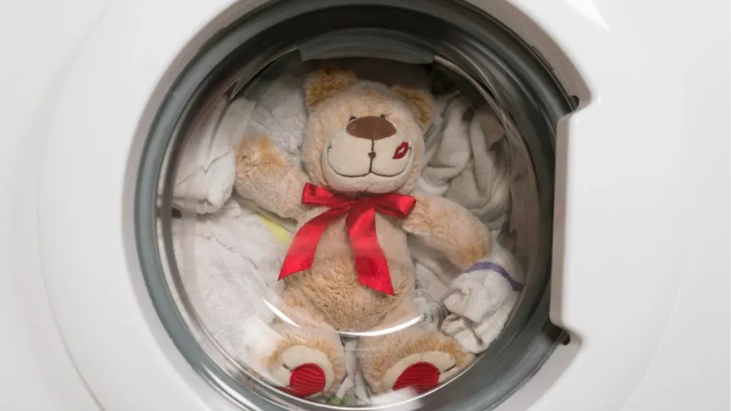 Brown teddy bear with a red bow being cleaned inside a front-load washing machine with towels for protection.