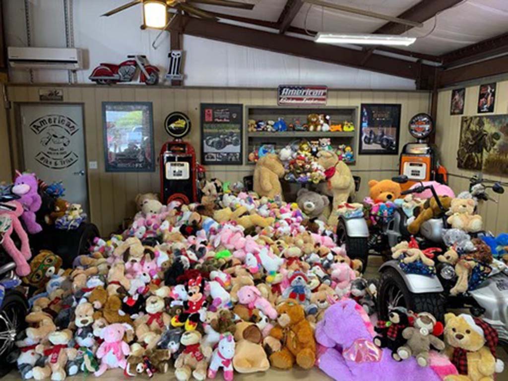 A room filled with hundreds of colorful plush toys and teddy bears, arranged around motorcycles and vintage decor, creating a cozy and nostalgic display.