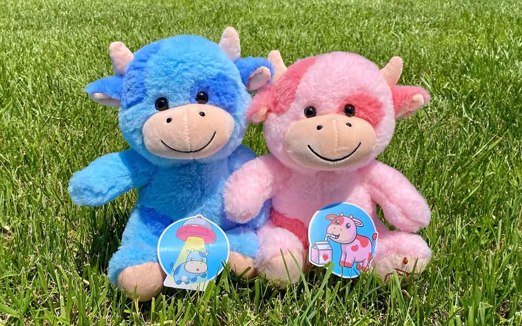 Two smiling plush cow toys, one blue and one pink, sitting on green grass with colorful cartoon tags attached to them.