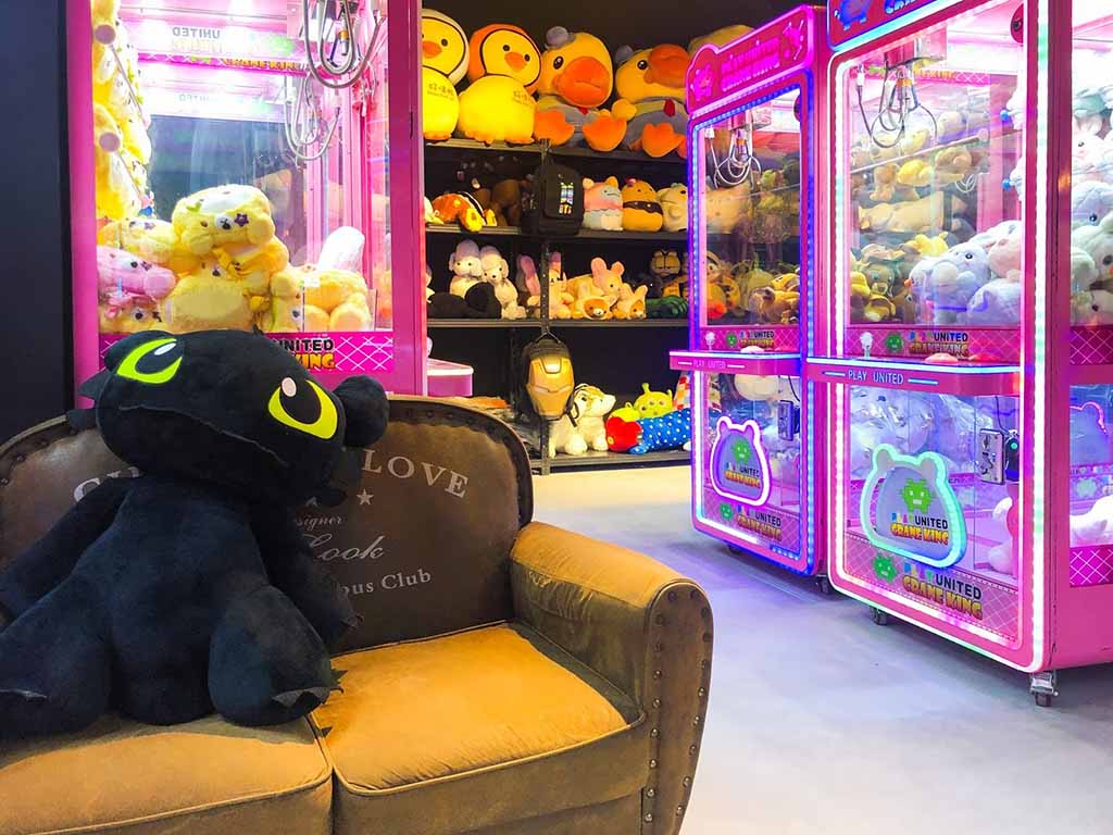 Colorful claw machines filled with plush toys beside a large black stuffed dragon on a sofa in a game arcade.