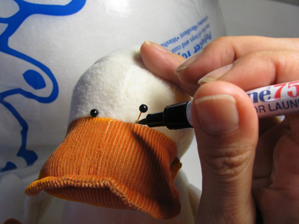 A craftsperson marking facial placement points with pins and a pen on a handmade plush duck with an orange corduroy beak.