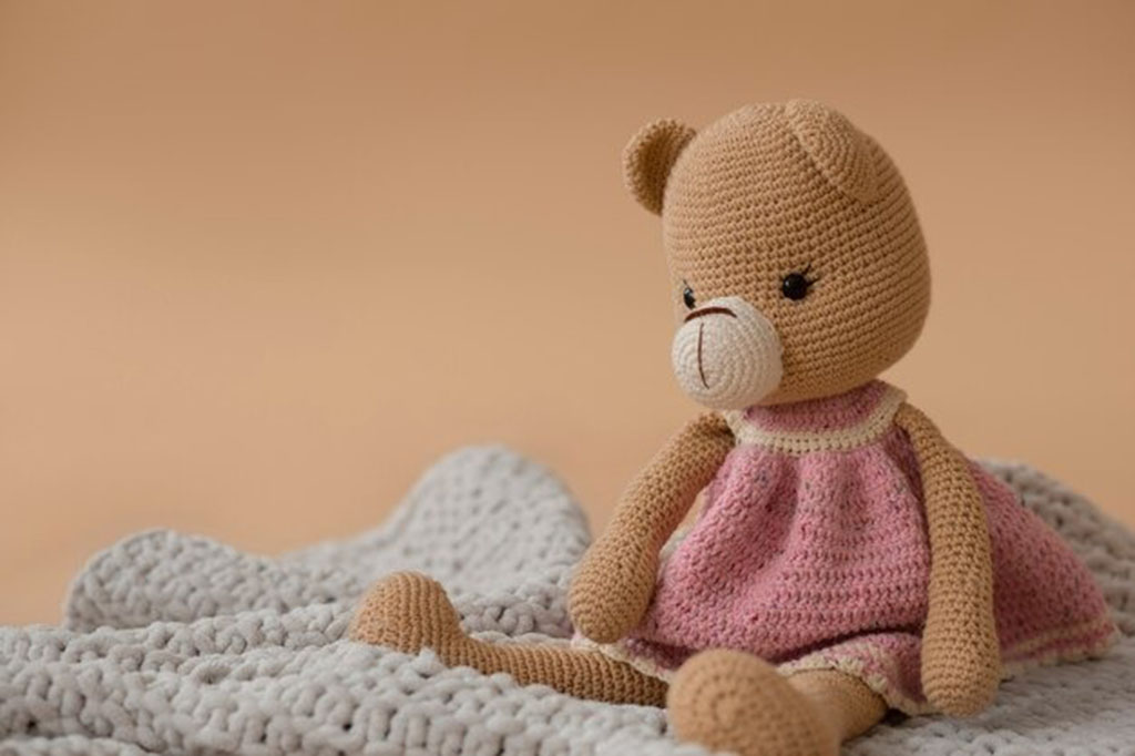 Handmade crochet teddy bear doll wearing a pink dress sitting on a soft knitted blanket.