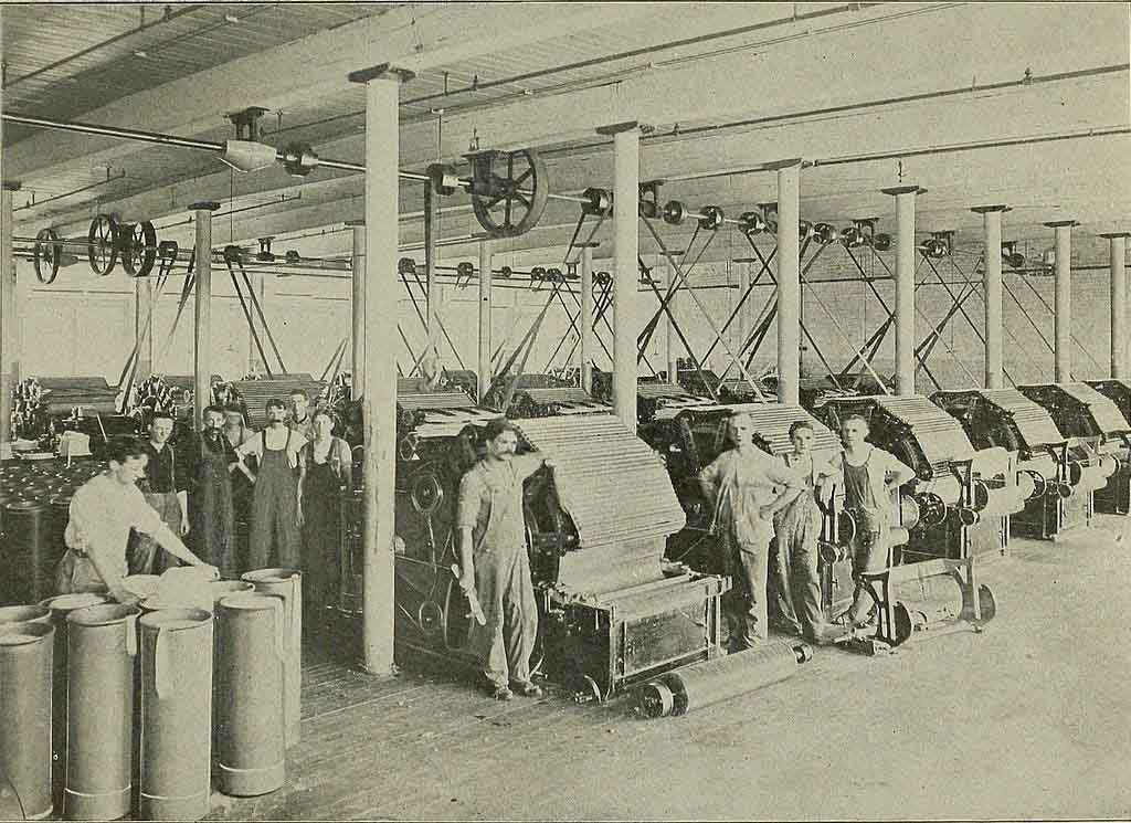 Vintage photo of workers operating large textile machines in an early 20th-century factory, showing the industrial production process with belts, pulleys, and rollers.