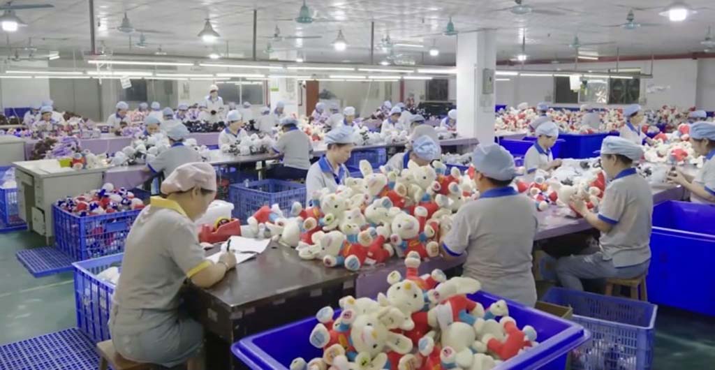 Factory workers in uniforms inspecting and assembling teddy bears at long tables with large blue bins filled with plush toys.
