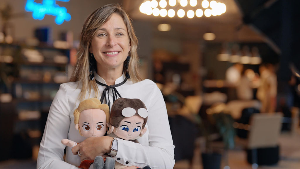 Smiling woman in a white blouse holding two custom plush dolls designed as animated characters, standing in a warmly lit office environment.