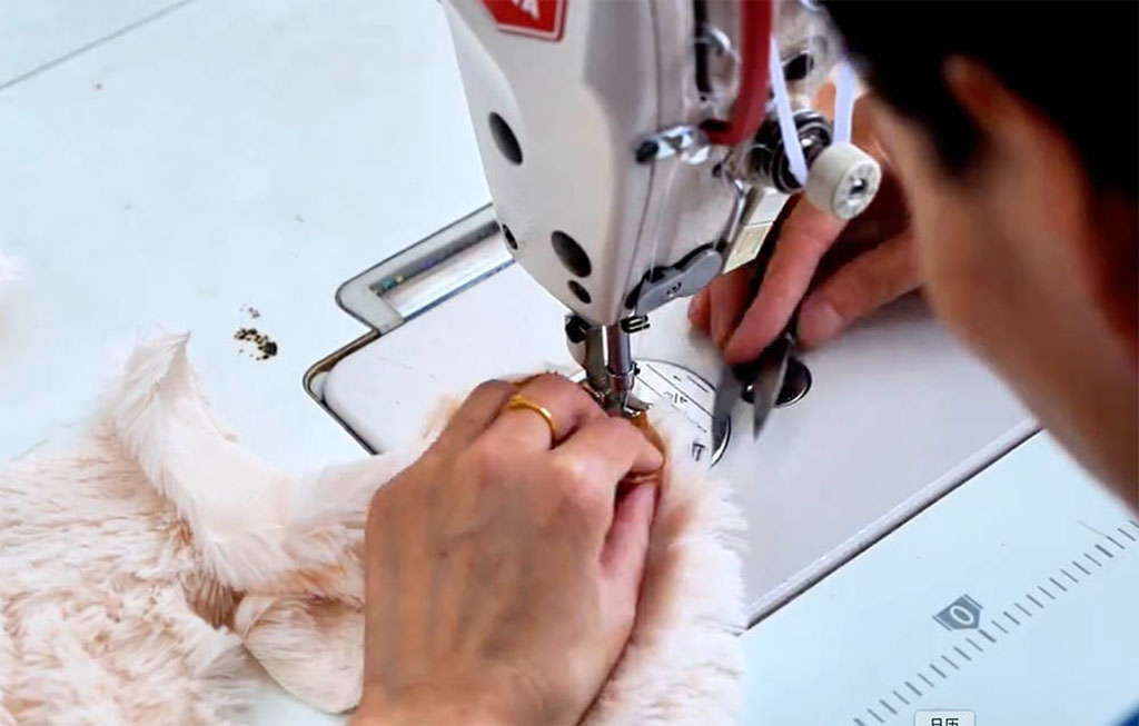 A craftsperson using a sewing machine to stitch soft plush fabric pieces together, part of the handmade plush toy production process.