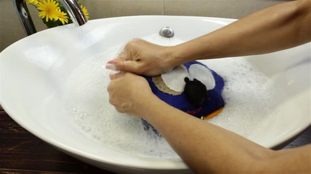 Person hand-washing a plush toy in a white sink filled with soapy water and bubbles.