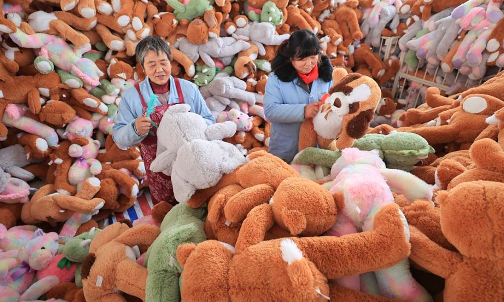 Two workers inspecting and grooming large plush toys in a factory filled with colorful stuffed animals ready for packaging.