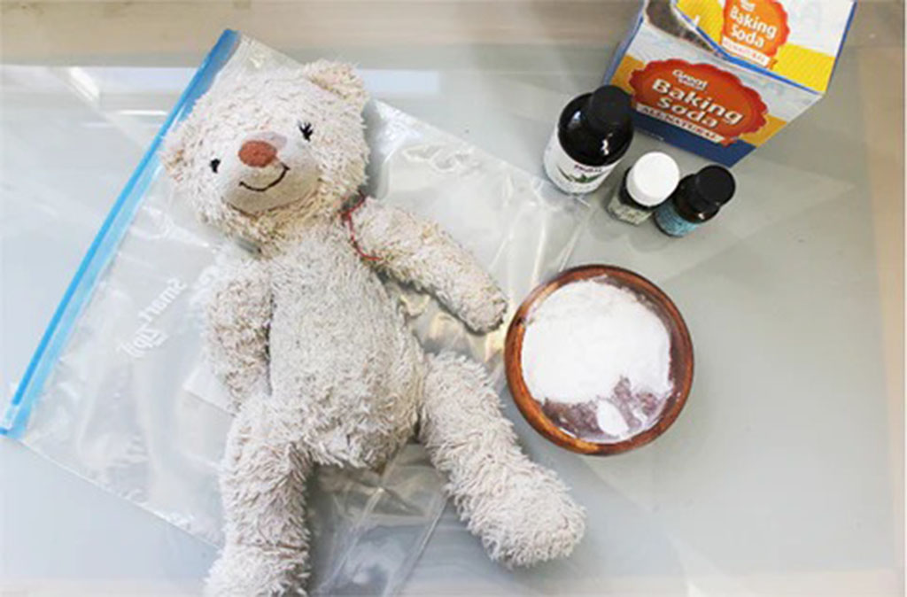 Worn teddy bear lying beside a bowl of baking soda, essential oil bottles, and a resealable plastic bag on a glass table, showing natural deodorizing supplies.
